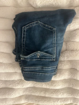 Mörkblå jeans med kontrastsömmar - Säljer ett par mörkblå jeans med vita kontrastsömmar och snygga detaljer på fickorna. Jeansen har klassisk femficksdesign och knappgylf. Materialet är slitstarkt denim som sitter snyggt och bekvämt.