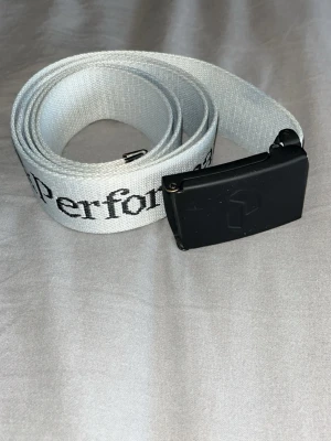 Vitt tygbälte från Peak Performance - Snyggt vitt tygbälte från Peak Performance med svart metallspänne och logga tryckt längs bandet. Perfekt accessoar för att lyfta din outfit med en sportig touch. Justerbar längd och klassisk design.