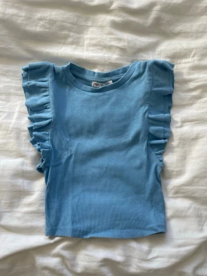 Blå croppad topp med volangärmar Zara - Supersöt ljusblå croppad topp från Zara med volangdetaljer på axlarna. Toppen är ribbad och har en tight passform som framhäver figuren. Perfekt för dig som gillar trendiga och feminina plagg.
