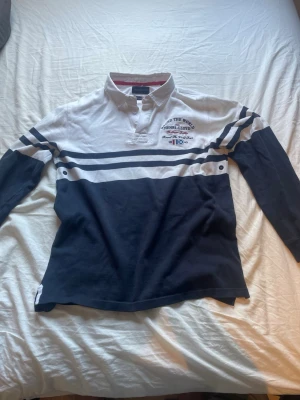 Randig långärmad Piké Henri Lloyd - Säljer en snygg långärmad skjorta från Henri Lloyd med marinblå och vita ränder. Skjortan har broderad logga och text på bröstet samt klassisk krage och knappar vid halsen. Tillverkad i bomull och perfekt för en sportig, avslappnad stil.