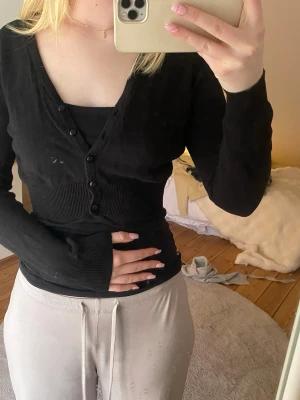 Svart kofta, kort - Säljer en svart, figurnära kofta med ribbad midja och långa ärmar. Koftan har V-ringning och knäppning framtill med fem svarta knappar. Perfekt att styla lager på lager för en clean och enkel look.