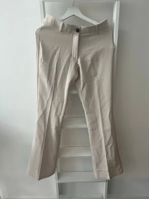Beige kostymbyxa - Stilrena beige kostymbyxor med raka/utsvängda ben. Byxorna har hällor för skärp, knapp och dragkedja framtill samt två passpoalfickor bak som ger en klassisk look. I storlek 164. Köparen står för eventuell frakt.