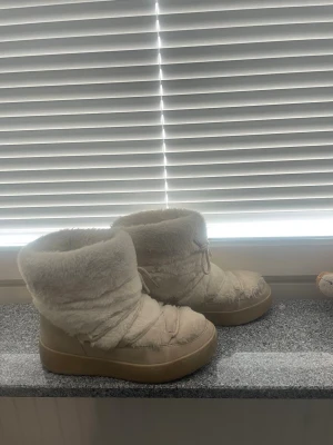 Beige fluffiga moon boots - Säljer ett par beige moon boots med fluffig, mjuk yta och högt skaft. Skorna har en chunky sula i gummi och snörning framtill för extra stilpoäng. Perfekta för kalla dagar när du vill hålla fötterna varma och se trendig ut. 