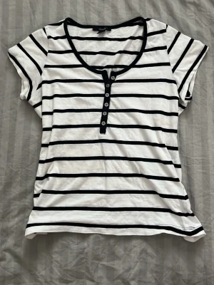 Randig t-shirt från Only med knappar - Säljer en vit och svart randig t-shirt från Only med korta ärmar och rund halsringning. T-shirten har dekorativa knappar framtill och är gjord i mjuk bomull. Perfekt för en avslappnad och stilren look.