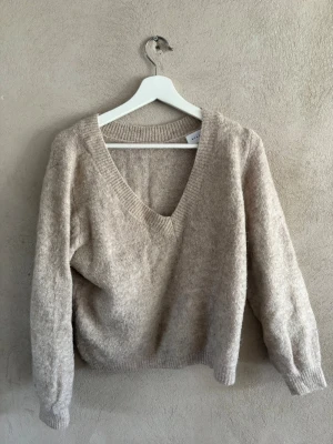 Beige stickad tröja med v-ringning - Mysig beige stickad tröja med djup v-ringning och långa ärmar. Tröjan har ribbade muddar vid ärmslut och nederkant, samt en avslappnad passform. Perfekt för lager på lager och enkel att matcha med olika stilar.
