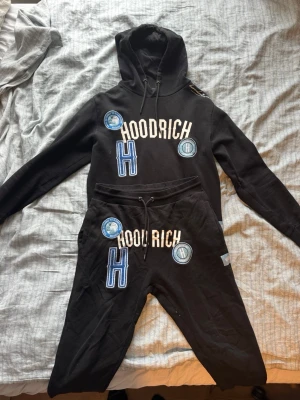 Svart Hoodrich set - Säljer ett svart set från Hoodrich med matchande hoodie och byxor. Setet har stora broderade patches och tryck i vitt, blått och ljusblått, samt texten 'HOODRICH' och bokstaven H på både tröja och byxor. Mjuk bomullskvalitet och avslappnad passform.