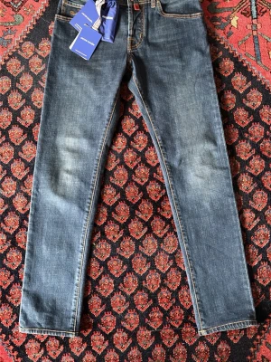 Blå jeans från Jacob Cohen - Säljer ett par klassiska blå jeans från Jacob Cohen med raka ben och snygga kontrastsömmar. Jeansen har fem fickor, normal passform och en diskret logopatch bak i midjan. Tillverkade i mjukt bomullsmaterial för skön känsla hela dagen.