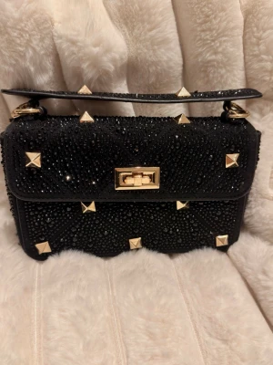 Svart handväska med nitar och glittrig - Säljer en svart handväska dekorerad med svarta strasstenar och guldiga fyrkantiga nitar. Väskan har ett kort handtag med guldfärgade detaljer och ett vridlås i guld. Perfekt för dig som gillar edgy och glamorös stil.