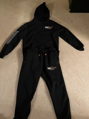 Svart Hoodrich set  - Säljer ett svart Hoodrich set med matchande hoodie och byxor. Setet har vita och orangea tryck med texten 'Hoodrich' på bröstet, ryggen, ärmen och byxan. Hoodien har huva och magficka, byxorna har dragsko i midjan. Perfekt för en avslappnad streetwear-look. Priset kan diskuteras.