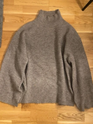 Grå ribbstickad polotröja - Mysig grå stickad polotröja med ribbad struktur och hög krage. Tröjan har en avslappnad passform och långa ärmar, perfekt för kyliga dagar. Materialet ser ut att vara en mjuk ull- eller ullmix som håller dig varm.