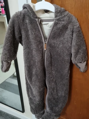 Brun fluffig morgonrock med huva - Mysig brun morgonrock i fluffigt material med huva och dragkedja framtill. Långa ärmar med muddar och helkroppsdesign som passar perfekt för att hålla värmen under kalla morgnar. Perfekt för att chilla hemma i maximal komfort.