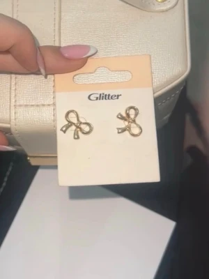 Guldiga rosettörhängen från Glitter - Säljer ett par söta örhängen i form av små rosetter från Glitter. De är guldfärgade och har en stilren, feminin design som passar perfekt till många olika looks. Örhängena är små och diskreta, vilket gör dem lätta att matcha med andra accessoarer.