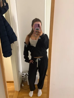 Svart mjukisset från victorias secret med huva - Säljer en svart hoodie från victorias secret med vit logga på bröstet och huva. Hoodien har dragkedja framtill och ribbade muddar vid ärmslut och nederkant. Perfekt för en avslappnad och cool look.