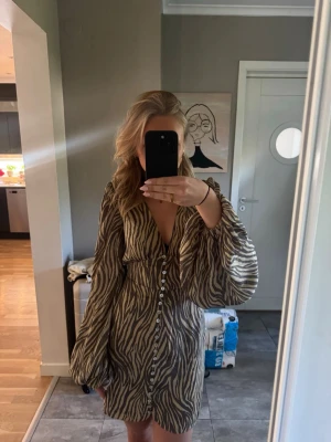 Zebramönstrad klänning Adoore - Snygg zebramönstrad klänning med v-ringning och långa ärmar. Klänningen har knappar framtill och är i ett mjukt, lätt material som ger en skön känsla. 