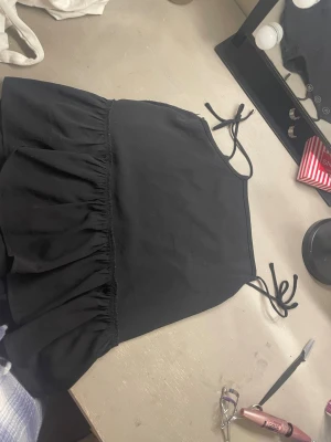 Svart linnetopp med volang och knyt - Säljer en svart linnetopp med tunna axelband som knyts upptill. Toppen har en volang nedtill som ger en luftig och söt look. Perfekt till jeans eller kjol för en chill stil.