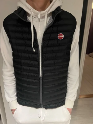 Svart dunväst från Colmar - Snygg svart dunväst från Colmar med quiltad design och dragkedja framtill. Västen har en diskret logga på bröstet och passar perfekt över en hoodie för en casual look. Lätt och smidig att bära under kyliga dagar. Liten defekt längst ner vid ryggen men fixad så gott det går. 