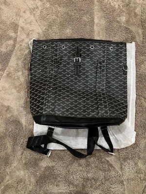 Svart ryggsäck med mönster - Snygg svart ryggsäck med det klassiska vita mönstret över hela väskan. Väskan har svarta läderdetaljer, justerbara axelband och en spänndetalj framtill. Perfekt för dig som vill ha en unik och trendig accessoar.