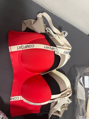 Röd och svart bh med breda band - Två bhar i rött och svart med breda, elastiska axelband där det står 'COMFORT'. Mjuka kupor utan bygel och slät design för extra komfort. Materialet ser ut att vara stretchigt och följsamt, perfekt för en bekväm känsla hela dagen.