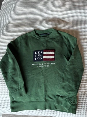 Lexington - Grön sweatshirt från Lexington - Grön sweatshirt från Lexington med broderad flagga och texten 'American Heritage Since 1993' på bröstet. Tröjan har rund hals, ribbade muddar och är tillverkad i mjuk bomull. Perfekt för en avslappnad och stilren look. Nypris ca 899kr, säljes för 375kr