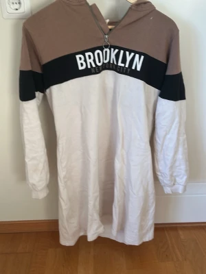 Brooklyn hoodie klänning  - Lång hoodie klänning  i vitt, brunt och svart med texten 'BROOKLYN NEW YORK CITY' på bröstet. Hoodien har huva, dragkedja framtill och långa ärmar. Tillverkad i mjukt bomullsliknande material och har en avslappnad passform.