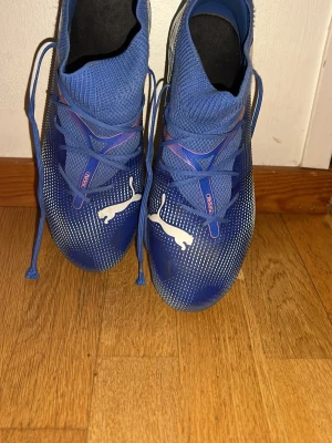 Blå Puma fotbollsskor med snörning - Säljer ett par blå fotbollsskor från Puma med högt skaft och snörning. Skorna har vita detaljer, inklusive den klassiska Puma-loggan på ovansidan. Ovandelen är tillverkad i ett syntetiskt material med stickad struktur för bra passform och kontroll.