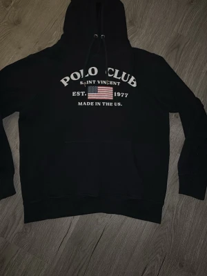 Marinblå hoodie Polo Club Saint Vincent - Svart hoodie från Polo Club Saint Vincent med tryck framtill som inkluderar amerikansk flagga och texten 'EST. 1977 MADE IN THE US’ Det är storlek S på denna och passar någon kring 165-173