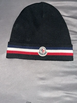 Svart Moncler mössa med randig kant - Säljer en svart stickad mössa från Moncler med klassisk ribbad kant i blått, vitt och rött. Mössan har Moncler-logga framtill och är tillverkad i mjukt material som sitter skönt på huvudet. Perfekt accessoar för kyliga dagar. den är ny inga problem med denna tar byten och den har nfc scan och pris kan alltid diskuteras 