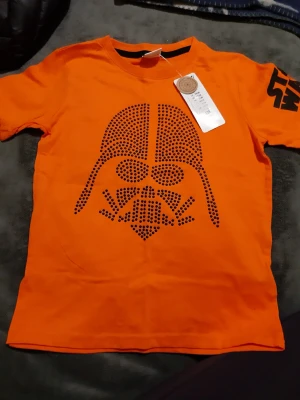 Helt ny. Orange Star Wars Darth Vader t-shirt - Helt ny med prislappen på. Cool orange t-shirt från Star Wars med Darth Vader-tryck i svart prickmönster på framsidan och stor Star Wars-logga på ärmen. Tillverkad i mjuk bomull och har en klassisk passform med korta ärmar. Perfekt för alla Star Wars-fans!
