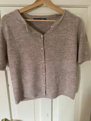 Beige kortärmad kofta med knappar - Mysig beige kofta med korta ärmar och v-ringning. Koftan har pärlemorskimrande knappar framtill och är stickad i ett mjukt material som ger en avslappnad look. Perfekt att slänga över en topp för en enkel men stilren outfit.