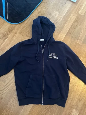 Mörkblå hoodie från Les Deux - Snygg mörkblå hoodie från Les Deux med dragkedja framtill och vit logga på bröstet. Hoodien har huva med dragsko och stora fickor framtill. Tröjan är aldrig använd. Storlek M. Köpt föt 1699kr säljer för 899kr. Inga skavander då ja aldrig användt den. Hör av dig vid frågor eller intresse!