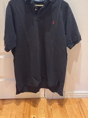 Svart pikétröja från Ralph Lauren - Klassisk svart pikétröja från Ralph Lauren med röd broderad logga på bröstet. Tröjan har korta ärmar, krage och knappar vid halsen. Tillverkad i bomull för en bekväm känsla och snygg passform.