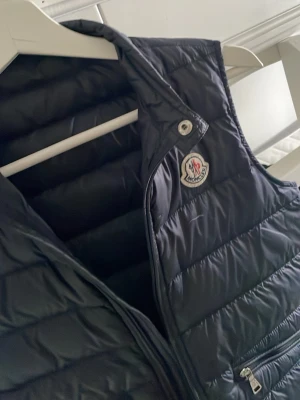 Svart dunväst från Moncler -  Stor i storlekn Snygg svart dunväst från Moncler med klassisk logga på bröstet. Västen har quiltad design, dragkedja framtill och praktiska fickor med dragkedja. Tillverkad i glansigt material som ger en exklusiv känsla. Perfekt för lager-på-lager och streetstyle.