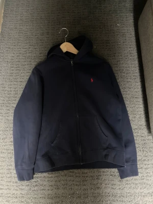 Mörkblå hoodie från Polo Ralph Lauren - Snygg mörkblå hoodie med dragkedja från Polo Ralph Lauren. Klassisk design med huva, kängurufickor och det ikoniska röda Polo-logot på bröstet. Tillverkad i mjuk bomullsblandning som känns skön mot huden. Perfekt för en avslappnad och stilren look. Köptes på kids brand store för ca 1 år sedan för 1195kr och mitt pris är 200kr nu👍 passar dig som är ungefär 165-175cm passar dessvärre inte mig längre då jag är 180 cm🙌