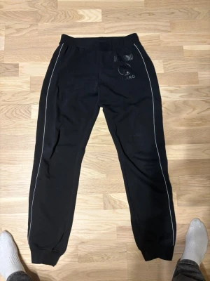 Svarta Moschino mjukisbyxor - Snygga svarta mjukisbyxor från Moschino med vita piping-detaljer längs benen och elastisk midja. Byxorna har Moschino Milano-logga tryckt framtill och är tillverkade i mjukt material som känns skönt mot huden. Perfekta för en avslappnad och trendig look.