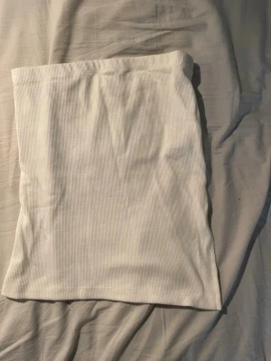 Vit ribbad tubtopp från Nelly - Säljer en enkel och stilren vit ribbad tubtopp från Nelly i storlek XS. Toppen är axelbandslös och har en mjuk, stretchig passform som sitter snyggt på kroppen. Perfekt att matcha med jeans eller kjol för en clean look.