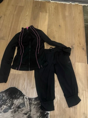 Svart träningsset med lila detaljer - Säljer ett svart träningsset med jacka och byxor. Jackan har dragkedja framtill och lila sömmar längs ärmar och sidor. Byxorna har resår i midjan och breda muddar vid bensluten. Perfekt för gym eller löpning.