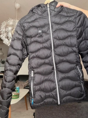 Svart dunjacka från decolt - Snygg svart dunjacka från decolt  med vågigt quiltat mönster och huva. Jackan har två dragkedjeförsedda fickor framtill och en silverfärgad dragkedja. Perfekt för kalla vinterdagar och riktigt lätt att bära. Jackan har en liten sminkfläck vid dragkedjan se bild. Men inget som inte går bort i tvätten. Är i stolek M men passar bättre som S