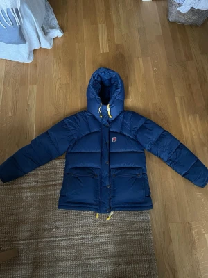 Mörkblå dunjacka fjällräven  - Snygg blå dunjacka från Fjällräven med stor huva och gula dragskor. Jackan har två stora fickor framtill med lock och tryckknappar samt en liten Fjällräven-logga på bröstet. Perfekt för kalla vinterdagar och riktigt varm.