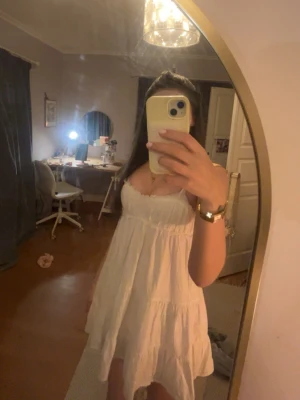 Vit kort sommarklänning med volang - Söt vit kortklänning med tunna axelband och volangdetaljer. Klänningen har en luftig passform och är perfekt för varma dagar. Tillverkad i ett lätt bomulls material som känns skönt mot huden. Enkel och stilren design som ger en fräsch look.