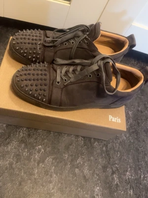 Svarta Louboutin sneakers med nitar - Säljer ett par svarta sneakers från Christian Louboutin med coola nitar på tån och klassisk röd sula. Skorna har svarta skosnören och är tillverkade i mocka med läderdetaljer på insidan. Perfekta för dig som vill sticka ut med din stil.