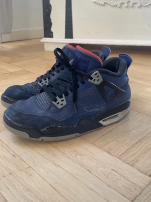 Blåa Air Jordan 4 sneakers - Snygga Air Jordan 4 sneakers i mörkblå mocka med svarta och grå detaljer. Skorna har meshpaneler på sidorna, svart snörning och en synlig luftbubbla i sulan. Insidan är röd och svart, och hälen pryds av den klassiska Jumpman-loggan.