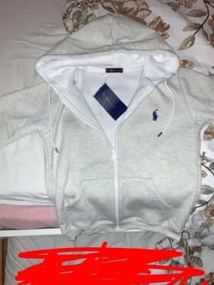 Vit hoodie med dragkedja från Polo Ralph Lauren - Snygg vit hoodie från Polo Ralph Lauren med dragkedja framtill och klassisk blå logga broderad på bröstet. Hoodien har huva med dragsko och två fickor framtill. Tillverkad i mjukt bomullsmaterial som känns skönt mot huden.