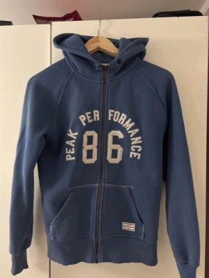 Blå hoodie Peak Performance 86 - Blå zip-hoodie från Peak Performance med trycket '86' och texten 'Peak Performance' i vitt på bröstet. Hoodien har huva med dragsko, två fickor fram och ribbade muddar. Perfekt för en avslappnad och sportig stil.