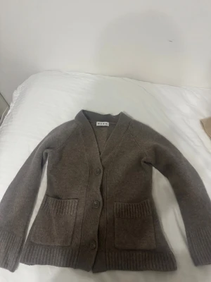 Brun kofta från Wera med fickor - Mysig brun kofta från Wera med tre knappar framtill och två stora fickor. Koftan har ribbade muddar vid ärmslut och nederkant samt en djup V-ringning. Perfekt för lager på lager och enkel att matcha.