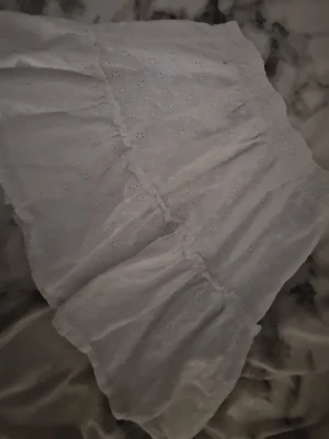 Vit volangkjol med broderade detaljer - Söt vit kortkjol med volanger och broderade små blommor. Kjolen har resår i midjan och är gjord i ett lätt och luftigt bomullstyg, perfekt för varmare dagar.