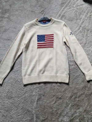 GANT Vit stickad tröja med USA-flagga  - Vit stickad tröja från Gant med en stor amerikansk flagga broderad på bröstet. Tröjan har rund halsringning, ribbstickade muddar och nederkant samt diskret Gant-brodyr på ena ärmen. Perfekt för dig som gillar klassisk och preppy stil.