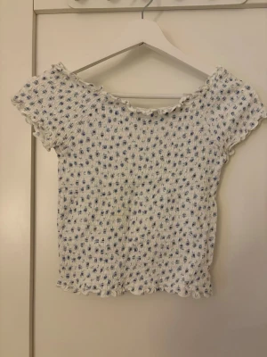 Vit blommig smockad topp - Supersöt vit topp med blått blommönster och smockad struktur. Toppen har korta ärmar och vågiga kanter vid hals, ärmslut och nederkant. Perfekt för en fräsch och somrig look!