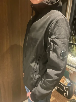 Svart hoodie från C.P. Company - Säljer en svart hoodie från C.P. Company med dragkedja framtill och huva. Hoodien har ribbade muddar, två sidofickor med dragkedja och en ikonisk linsdetalj på ärmen. Tillverkad i ett mjukt och slitstarkt material som passar perfekt till streetwear-stilen.