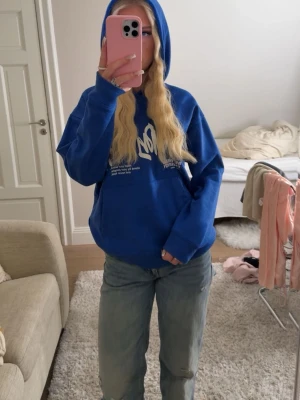 Blå hoodie med vit text - Snygg blå hoodie med vit grafisk text på bröstet . Hoodien har huva, magficka och ribbade muddar. Perfekt för en chill och avslappnad stil. Materialet är mjukt och skönt, troligen bomullsblandning.