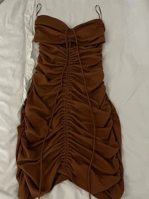 Brun omlott kortklänning med snörning - Snygg brun kort klänning med tunna axelband och trendig snörning framtill. Klänningen har omlottdetaljer och rynkade partier som ger en cool och modern look. Perfekt för dig som vill sticka ut med en unik och stilren outfit.